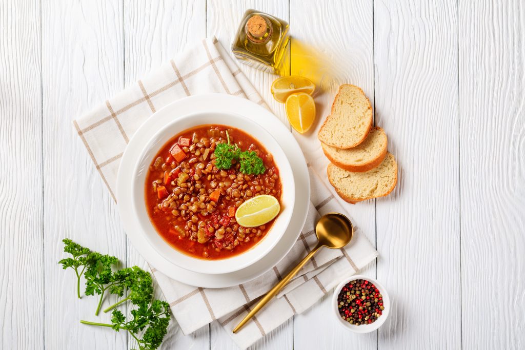 Curried Lentil Chickpea Stew, Lentil soup with vegetables and lemon wedges in a white bowl on a wooden table with a golden spoon and toasts, horizontal view from above, flatlay, free space Curried Lentil Chickpea Stew, Lentil soup with vegetables and lemon wedges in a white bowl on a wooden table with a golden spoon and toasts, horizontal view from above, flatlay, free space